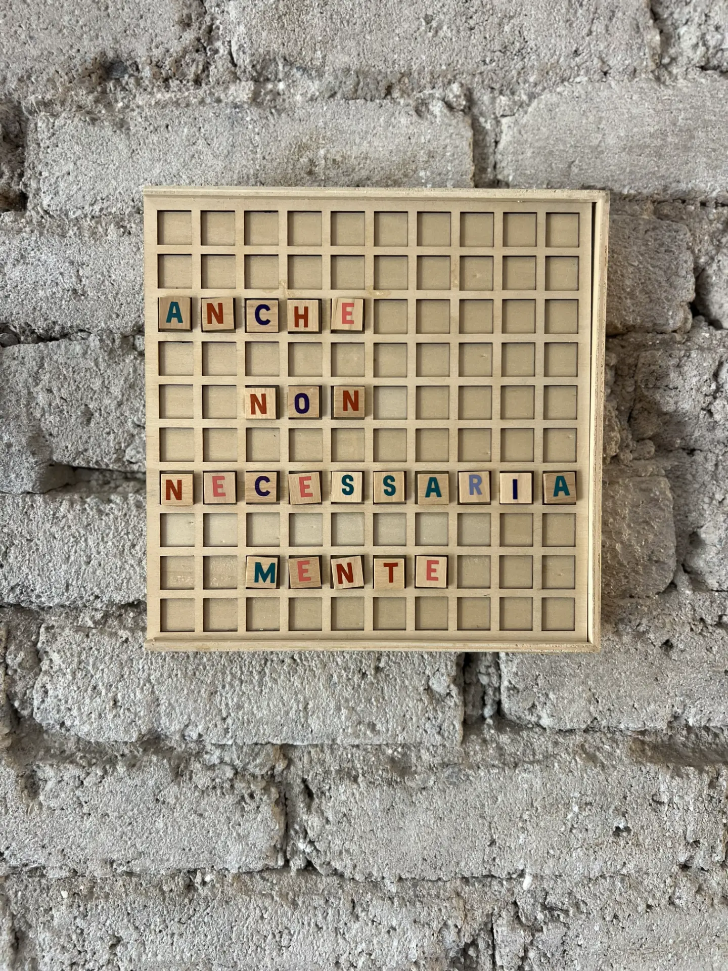 Bacheca quadrata in legno chiaro con griglia di scomparti, appesa a una parete di mattoni grigio chiaro dalla texture ruvida. Tessere di legno con lettere colorate compongono la frase «Anche non necessariamente».